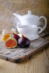 A white cup and a white teapot with tea, a jar of honey and fresh ripe figs on the cutting board on the table.