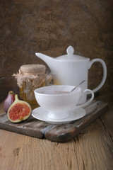 A white cup and a white teapot with tea, a jar of honey and fresh ripe figs on the cutting board on the table.