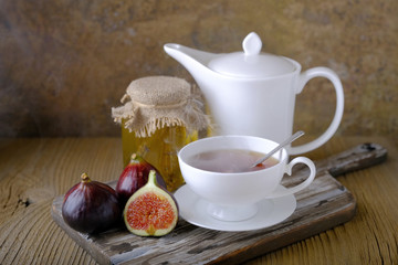 A white cup and a white teapot with tea, a jar of honey and fresh ripe figs on the cutting board on the table.