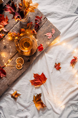 Cozy autumn or winter at home. A cup of tea, autumn casts a book a garland on a wooden table near a bed with warm plaids. Lifestyle autumn hygge lagom?concept of a holiday and autumn weekend.Flat lay