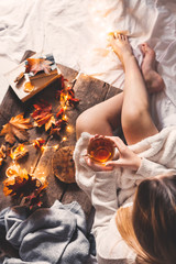 Cozy autumn or winter at home. Girl with a cup of tea, autumn casting book a garland on a wooden table near the bed with warm plaids. Lifestyle autumn lagom concept of relaxation and autumn weekend.