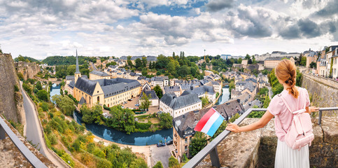 A happy Asian traveller girl holds the flag of Luxembourg and admires the great panoramic view of the Grund town from the viewing spot. Tourism, recreation and life in the country concept - obrazy, fototapety, plakaty