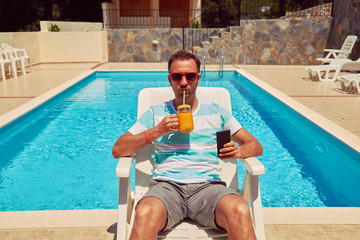 Attractive man using cellphone while drinking juice near swimming pool.