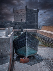 Fantasy view of old traditional wooden blue fisherman boats. Boats standing on the dock in the old city harbour. Stonewall and stone tower in the background. Essaouira, Morocco.