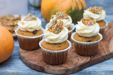 Pumpkin cupcakes with cream cheese frosting decorated with pecan nuts, maple syrup, caramel bites and chocolate toffee candies on wooden rustic background
