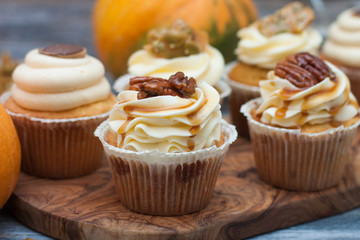 Pumpkin cupcakes with cream cheese frosting decorated with pecan nuts, maple syrup, caramel bites and chocolate toffee candies on wooden rustic background