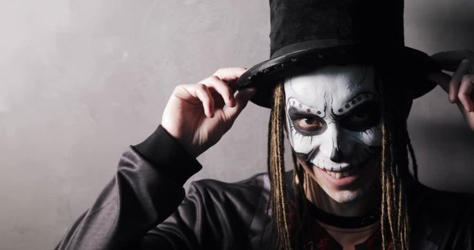 Guy with scary makeup in black hat and dreads hairs. Portrait of man in witcher costume looking at camera and smiling viciously on grey background. Celebrating Halloween holiday concept. Ghost freak.