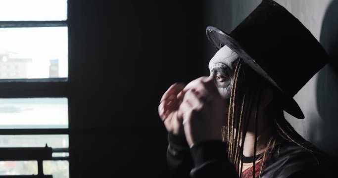 Ghost gothic freak on grey wall background near window. Portrait of man in witcher costume and black hat for Halloween with dreads hairs, side view. Scary makeup of black and white on his face.