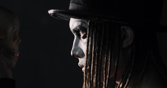 Young guy in carnival costume of witcher or ghost in black hat with dreads hairs on dark background. Portrait of man with scary gothic makeup for Halloween looking at skull in his hand, side view.