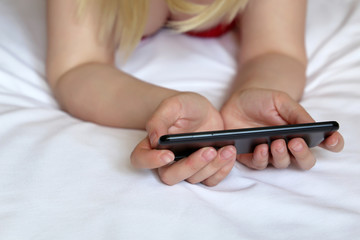 Blonde girl with smartphone, woman using mobile phone laying on the bed. Concept of online addiction, social media, sms messages
