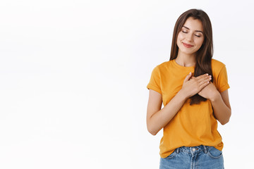 Tender, carefree romantic, dreamy young brunette woman in yellow t-shirt, holding hands pressed to heart, filled with warmth of love and passion, close eyes and smiling, recall happy moment