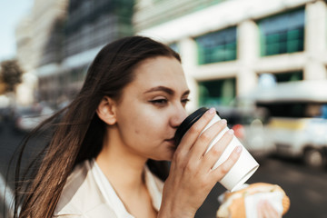 Young business girl in the beige coat eats on the go, holding a roll in her right hand and a paper cup in her left hand, rushing to a meeting