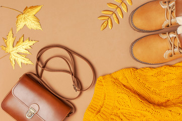 Brown leather women bag, orange knitted sweater, warm boots, golden autumn leaf on brown background top view flat lay. Fashionable women's accessories. Autumn Fashion Concept. Stylish Lady Clothes