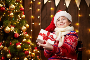 Cheerful santa helper girl with gift box sitting indoor near decorated xmas tree with lights, dressed in red sweater - Merry Christmas and Happy Holidays!