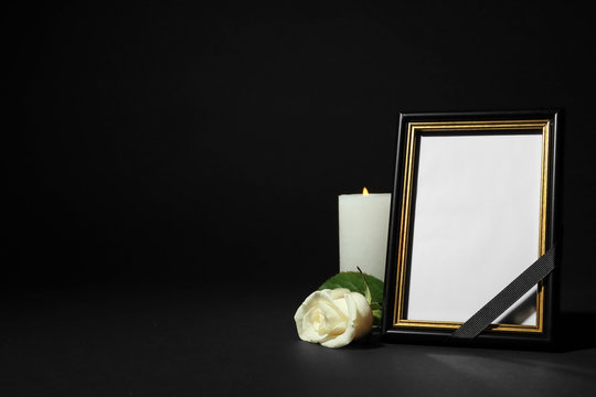 Funeral photo frame with ribbon, white rose and candle on dark table against black background. Space for design