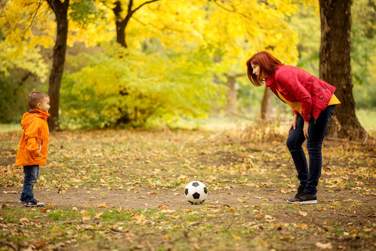 Little Boy Playing Football With Mother In Autumn Park. Mom Plays Soccer With Son Outdoors: Woman Leaned Forward Ready To Rush In Game, Ball Is Laying Between Them. Yellow Trees In Background