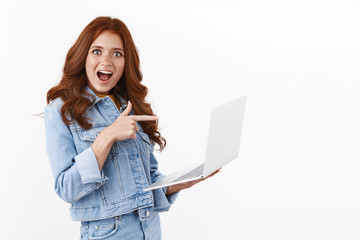 Amused enthusiastic redhead female in denim jacket, standing half-turned over white background, hold laptop pointing finger computer screen, checking out awesome offer internet, smiling astonished