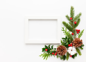 Christmas composition  with photo frame, cotton flower, branches of spruce and holly with red berries on white background. Merry christmas greeting card with empty space for holiday text.