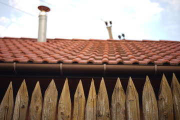 Fence made of sharp wooden stakes. Background house with red tiled roof and blue sky. Blurred