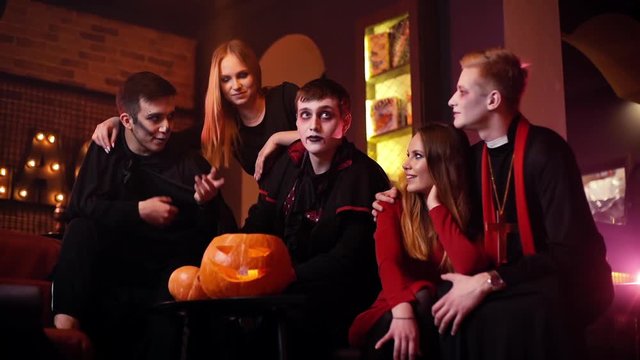 Young man in festive priest costume hugs his girlfriend at Halloween meeting of friends in cafe. Guy in the image of Count Dracula tells stories. Holiday carved pumpkin on table. Shot in slow motion