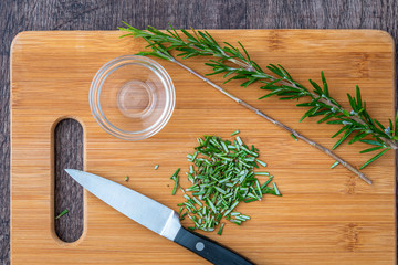 Fresh sprigs of rosemary with on a bamboo cutting board, chopped rosemary, paring knife and small glass bowl
