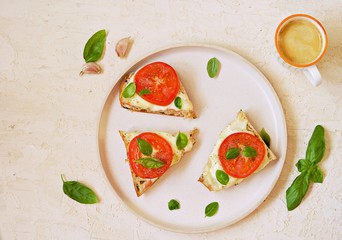 Open hot sandwiches with mozzarella cheese, fresh tomato and basil on a white plate on a light concrete background. Italian food. Top view, copy space.