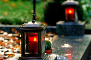old metal grave lights with burning candles on a grave with autumn leaves in the background