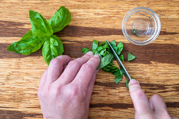 Woman’s hands chopping fresh basil with a paring knife on a rustic grove bamboo cutting board,...