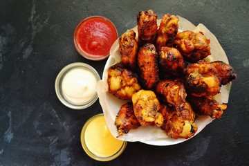 Fried chicken wings. Chicken wings on a dark background on a blackboard. Barbecue meat, grill. Sauce in bowls, ketchup, mayonnaise, mustard, cheese sauce. Appetizing fried meat. Place for your text