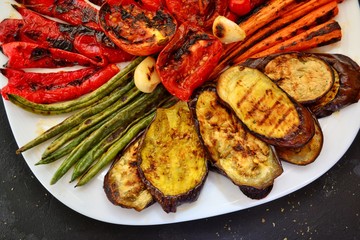 Grilled vegetables on a white plate. Appetizing grilled vegetables in a plate on a dark background, close-up. Vegetarian wholesome food. Baked eggplant, bell peppers, tomatoes, asparagus and carrots.