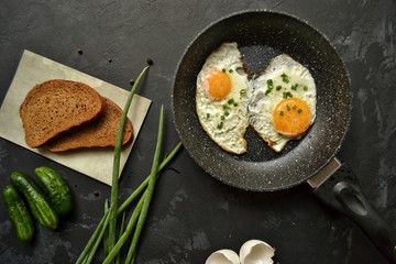 Tasty breakfast. Food on the table. Food on a dark concrete background. Fried eggs in a pan. Eggs, green onions, brown bread, cucumbers.