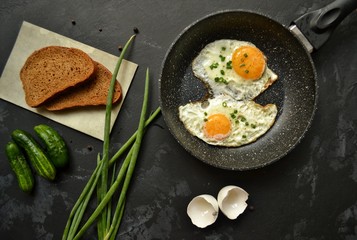 Tasty breakfast. Food on the table. Food on a dark concrete background. Fried eggs in a pan. Eggs, green onions, brown bread, cucumbers.
