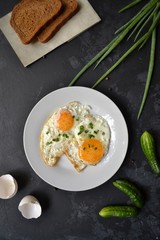 Tasty breakfast. Food on the table. Food on a black concrete decorative background. Fried eggs in a white plate. Eggs, green onions, brown bread, cucumbers.