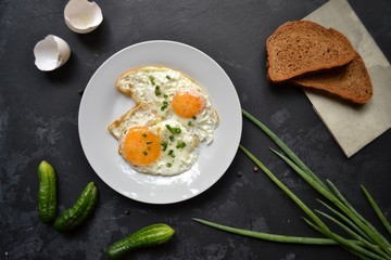 Tasty breakfast. Food on the table. Food on a black concrete decorative background. Fried eggs in a white plate. Eggs, green onions, brown bread, cucumbers.