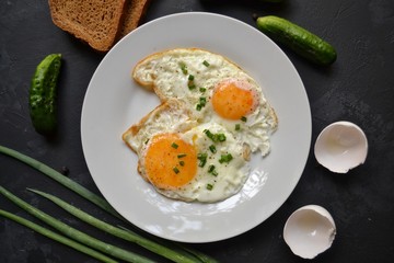 Tasty breakfast. Food on the table. Food on a black concrete decorative background. Fried eggs in a white plate. Eggs, green onions, brown bread, cucumbers.