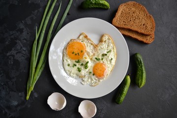 Tasty breakfast. Food on the table. Food on a black concrete decorative background. Fried eggs in a white plate. Eggs, green onions, brown bread, cucumbers.