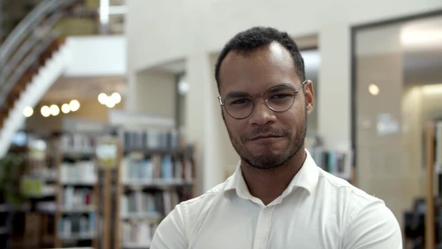Smiling African American man posing at public library. Front view of confident young guy standing in front of bookshelves. Concept of confidence