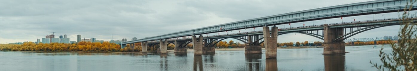 Panoramic shot of river Ob and its bank in Novosibirsk, Russia during autumn: a metro bridge and transport bridge on the right, the shoreline with yellowed fall trees and cityscape in the background