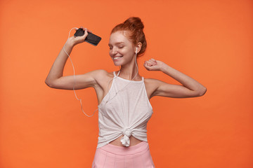 Studio photo of charming young woman wearing her red hair in knot posing over orange background, enjoying music track with closed eyes, dancing with raised hands