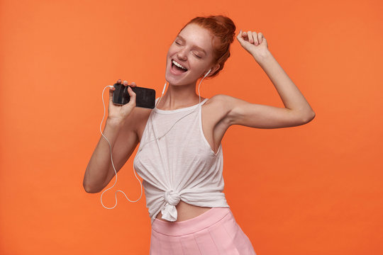Portrait of attractive young readhead woman with foxy hair in knot listaning to music with earphones, wearing white top and pink skirt, dansing over orange background with closed eyes