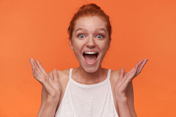 Indoor photo of joyful young readhead female with bun hairstyle wearing white top, looking to camera with surprised face and wide mouth opened, posing over orange background with raised palms
