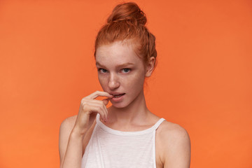 Studio shot of flirty young female lady in white top wearing her foxy hair in knot, looking at camera and keeping forefinger on underlip, standing over orange background