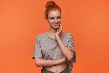 Positive attractive young readhead female with bun hairstyle smiling broadly and demonstrating her white perfect teeth, touching face with raised hand and looking to camera positively