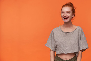Indoor photo of happy young readhead lady with bun hairstyle looking asidewith joyful smile, standing over orange background in grey t-shirt and green shorts. Positive human emotion facial expression