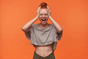 Indoor photo of stressed young woman wearing her foxy hair in knot isolated over orange background, covering ears with hands to avoid annoying sounds, screaming loud with wide mouth opened
