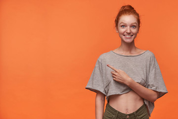 Obraz premium Studio shot of surprised readhead young lady with bun hairstyle pointing aside with forefinger, smiling joyfully to camera with wide eyes opened, isolated over orange background