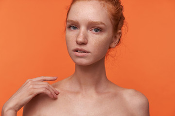 Indoor shot of attractive young female wearing her red hair in bun hairstyle standing over orange background, looking at camera gently with ajar mouth and touching shoulder with raised hand