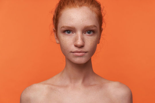 Studio photo of beautiful young readhead female with casual hairstyle standing over orange background without clothes, looking at camera with calm face