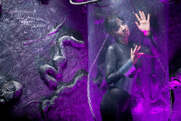 Shot of a futuristic young person posing near glass space capsule with wires and purple neon light on black background
