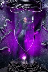 Shot of a futuristic young person posing near glass space capsule with wires and purple neon light on black background